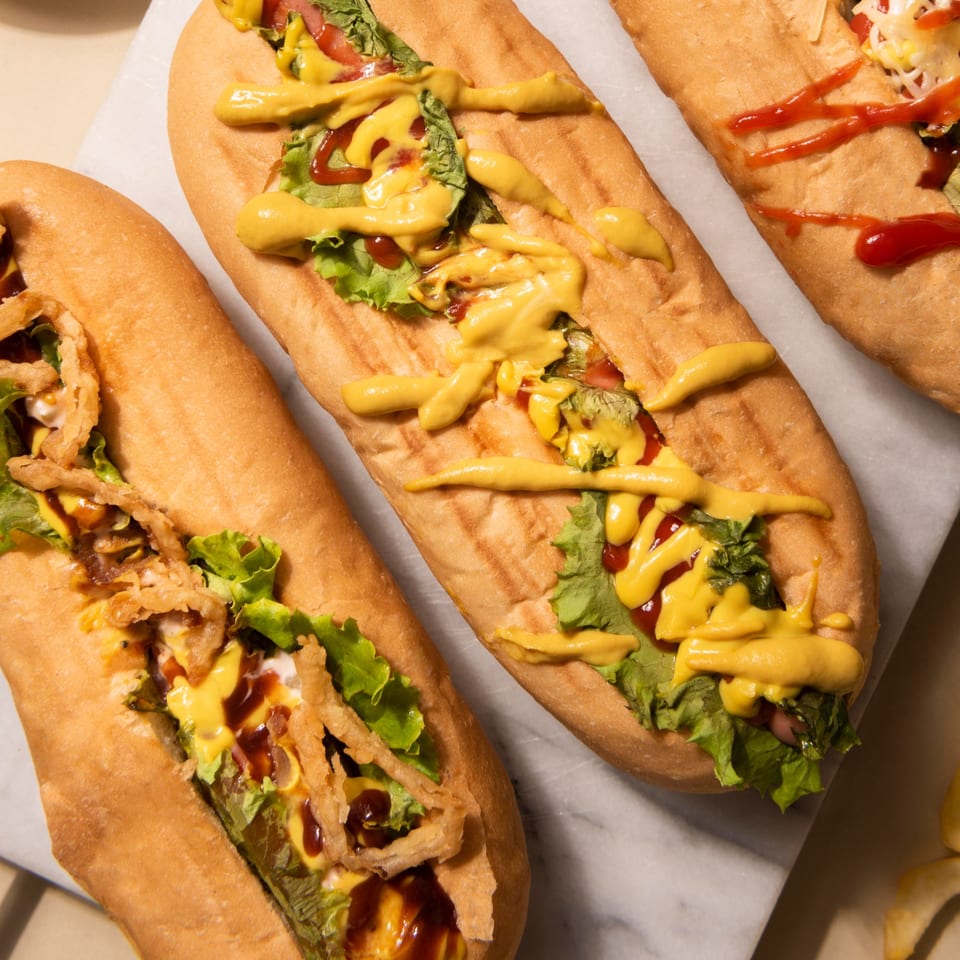 Three hot dogs in buns, topped with lettuce, mustard, ketchup, and crispy onions on a marble surface.
